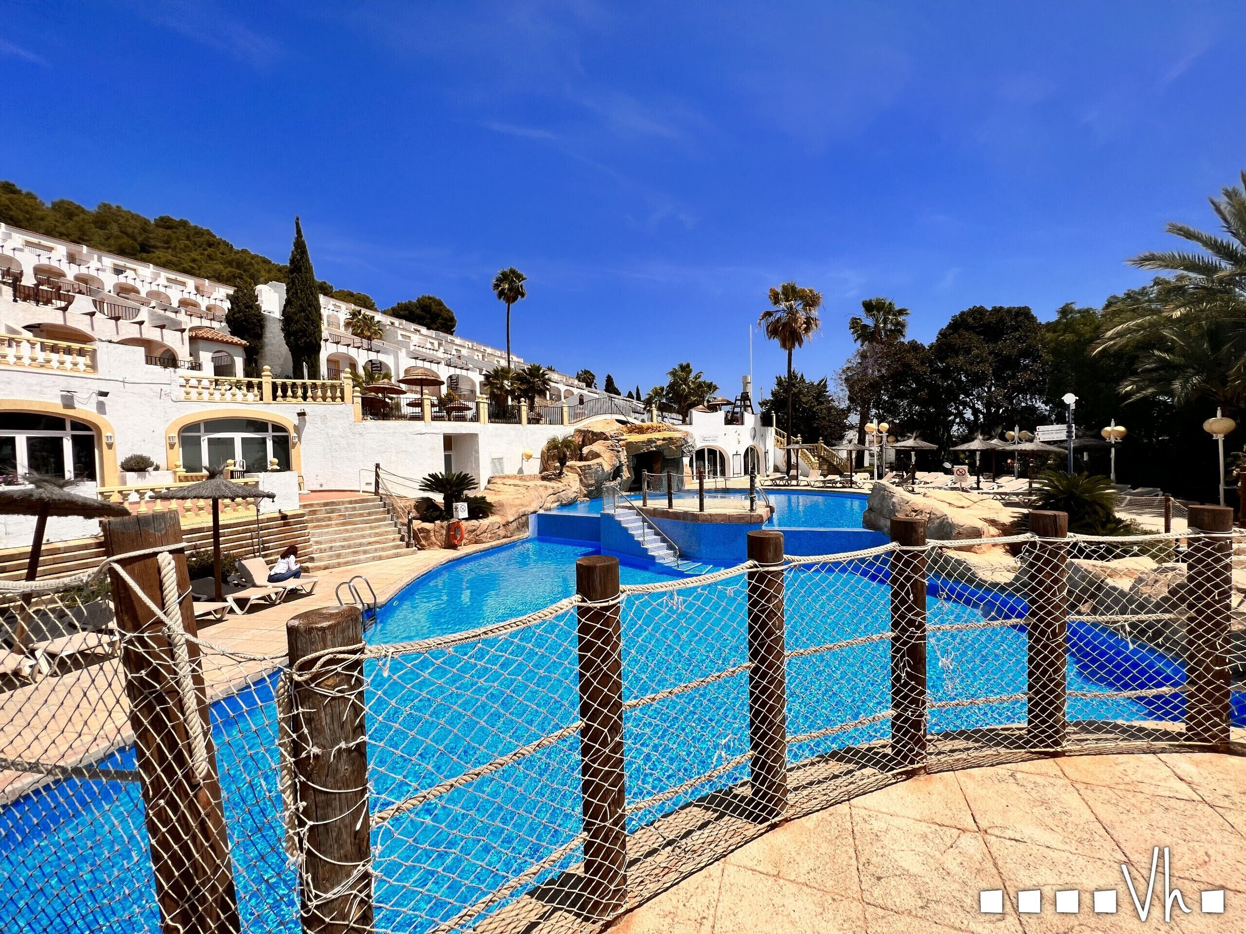 A pool area with a net fence, lounge chairs, and a building with balconies in the background.