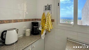 Cucina con macchina da caffè, bollitore e contenitore per il tè sul piano di lavoro, una finestra con vista sul mare e un porta asciugamani con asciugamani gialli.
