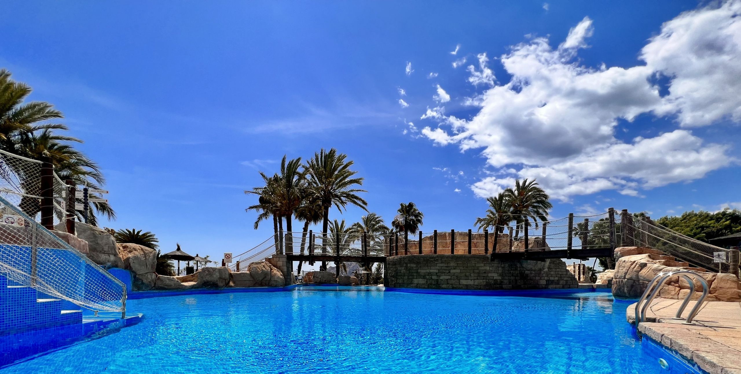 Una grande piscina esterna con acqua cristallina e azzurra, circondata da palme e un ponte in pietra.