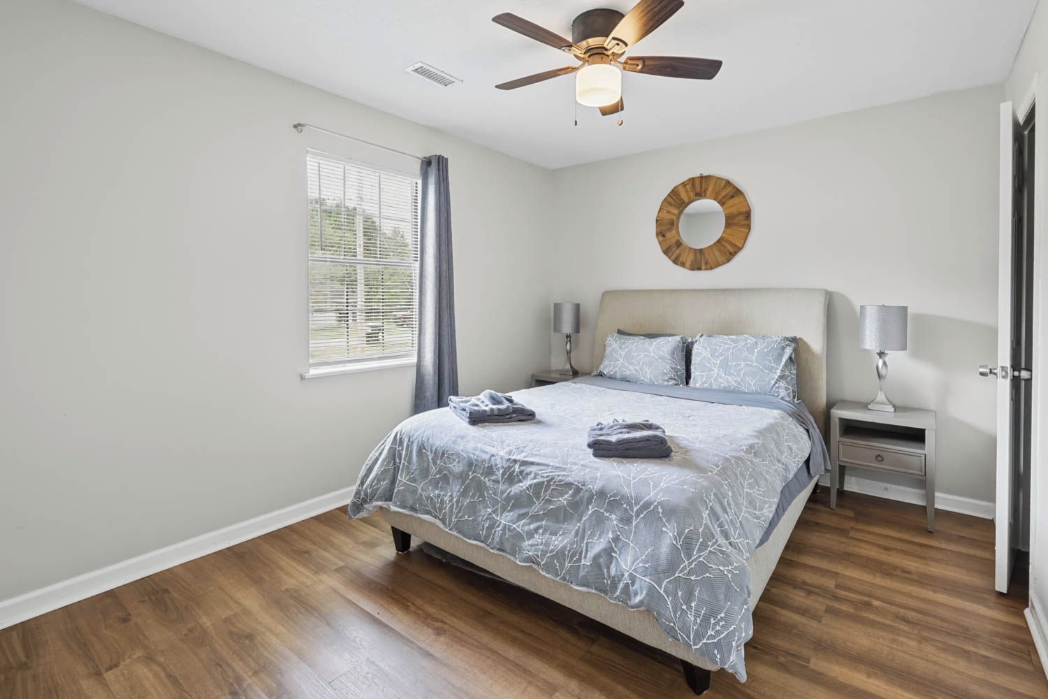 A bedroom with a bed, a ceiling fan, a bedside table with a lamp, a window with blinds, and a wall-mounted mirror.