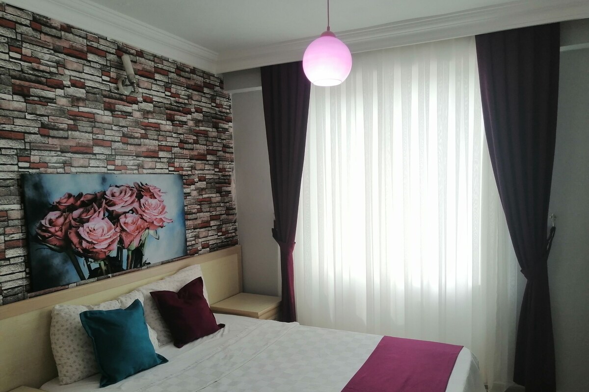 A hotel room with a brick-patterned wall, a painting of pink roses, a bed with a white and purple bedspread, and a pink pendant light.