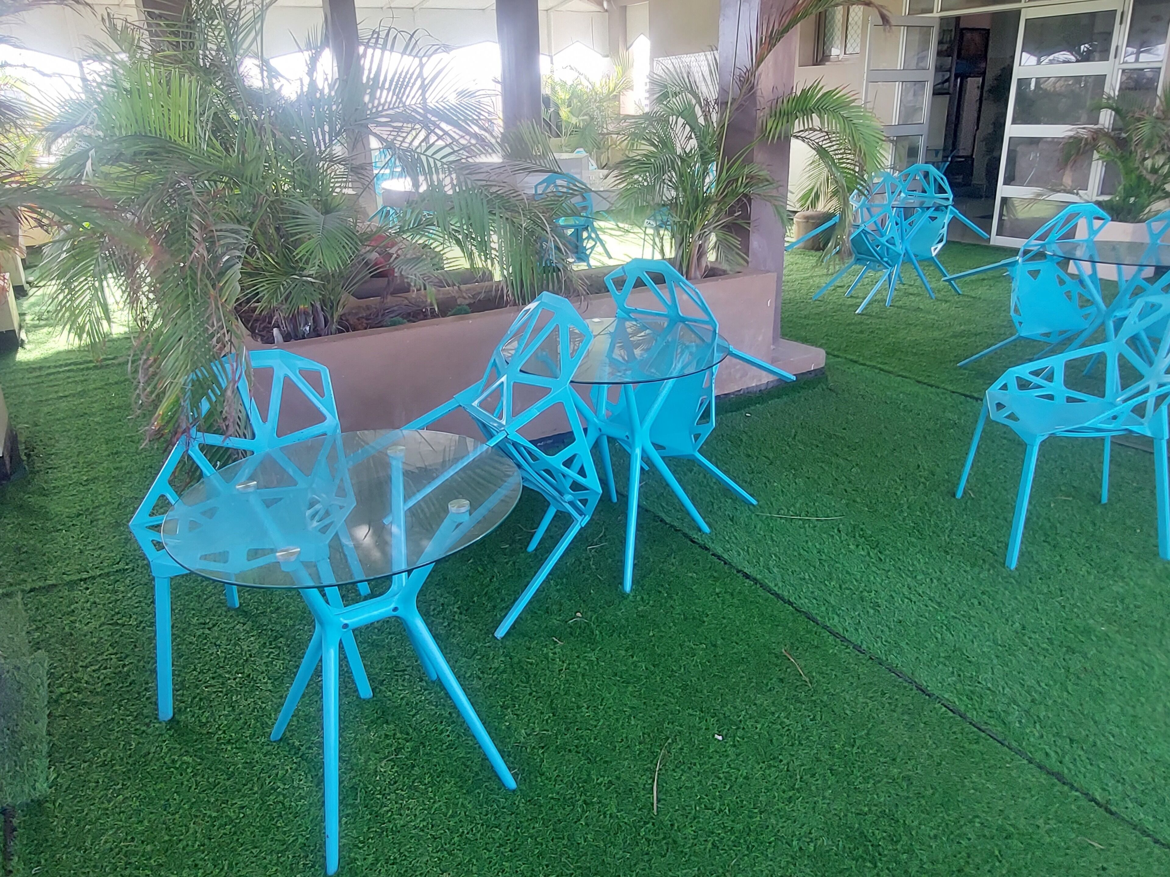 Outdoor seating with blue chairs and a glass table, surrounded by artificial grass and palm trees.