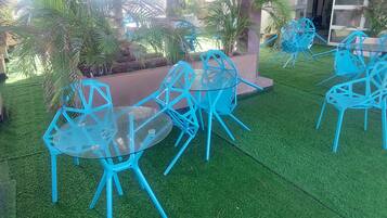Outdoor seating with blue chairs and a glass table, surrounded by artificial grass and palm trees.