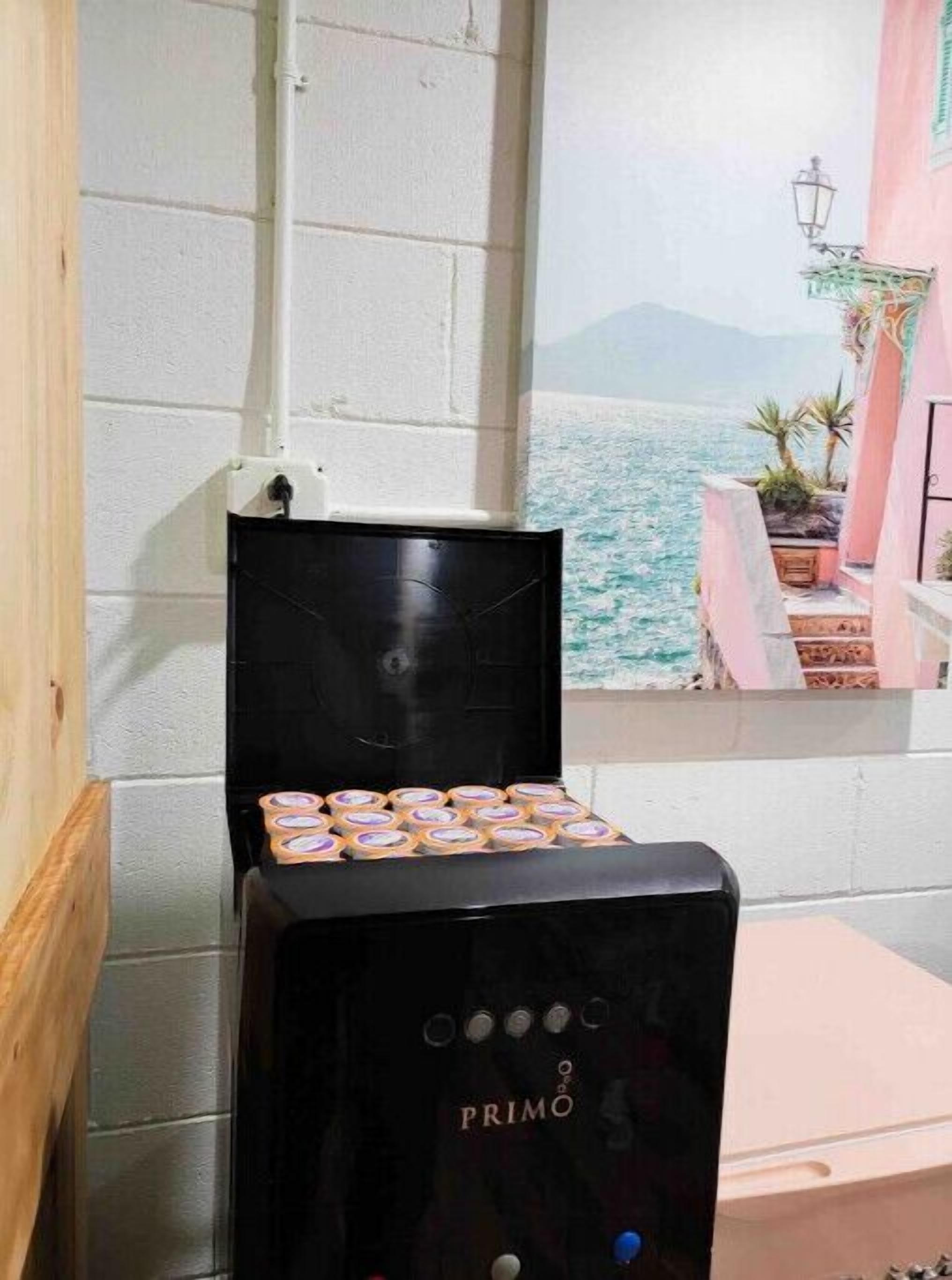 A black espresso machine with the brand name PRIMO, placed on a wooden floor next to a white wall.