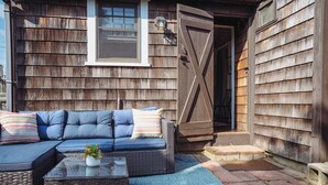 A cozy outdoor seating area with a sectional sofa and a coffee table, set against a wooden exterior wall with a large barn-style door. - Steps to a private beach in a classic Cape cottage. Pet-friendly! (Dennis)