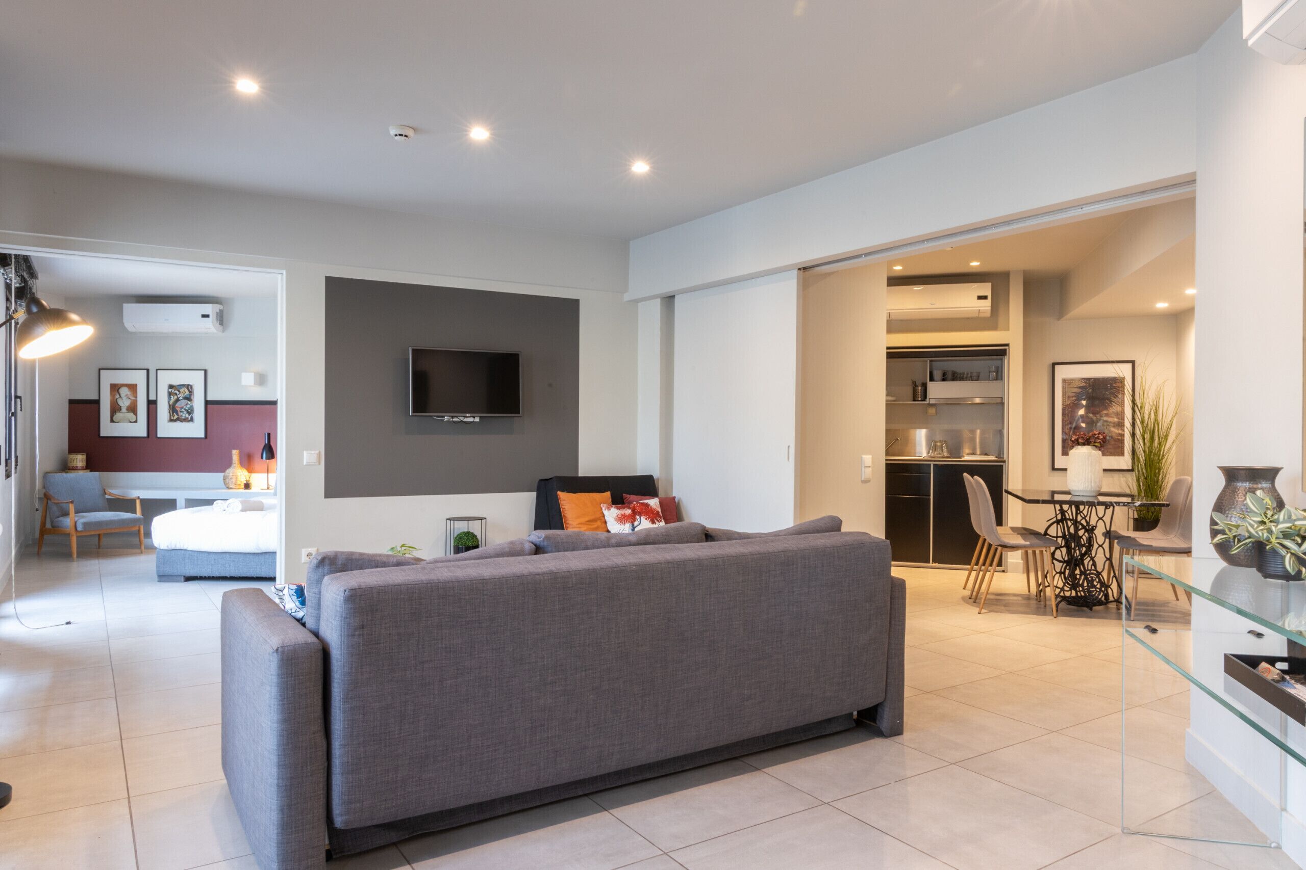 Un salon moderne avec un canapé gris, un téléviseur à écran plat fixé au mur et un coin repas comprenant une table et des chaises en verre.
