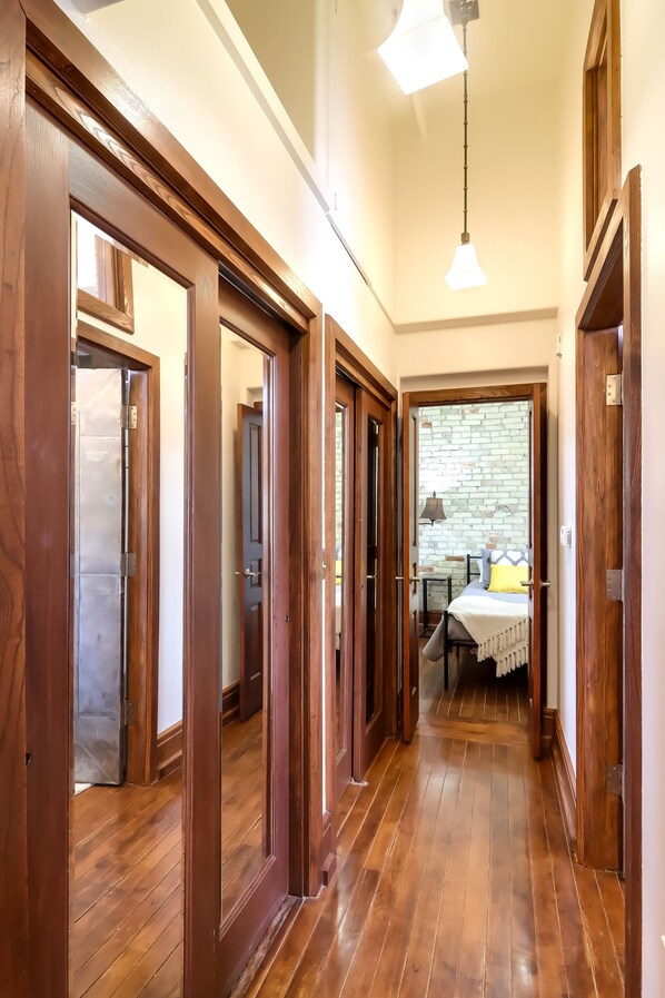Um corredor com piso de madeira e portas, que leva a um quarto com parede de tijolos e uma cama com travesseiro amarelo.