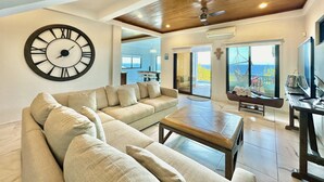 A spacious living room with a large sectional sofa, a coffee table, and a wall-mounted clock. There is a large window offering a view of the ocean.