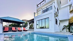 A swimming pool with red lounge chairs, a covered seating area, and a modern building with large windows and a staircase.