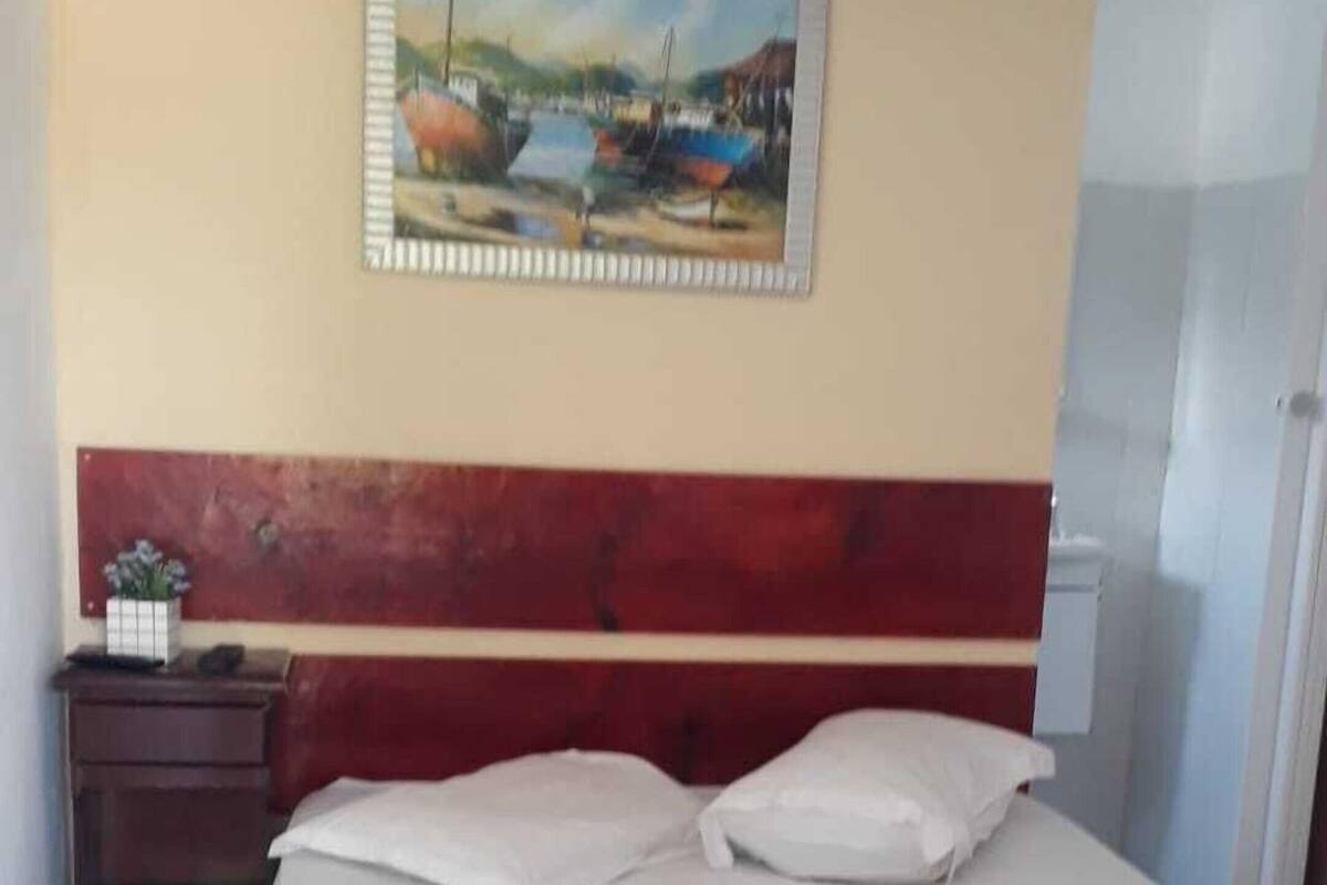 A neatly made bed with white and black bedding, a painting of a coastal scene on the wall, and a red headboard.