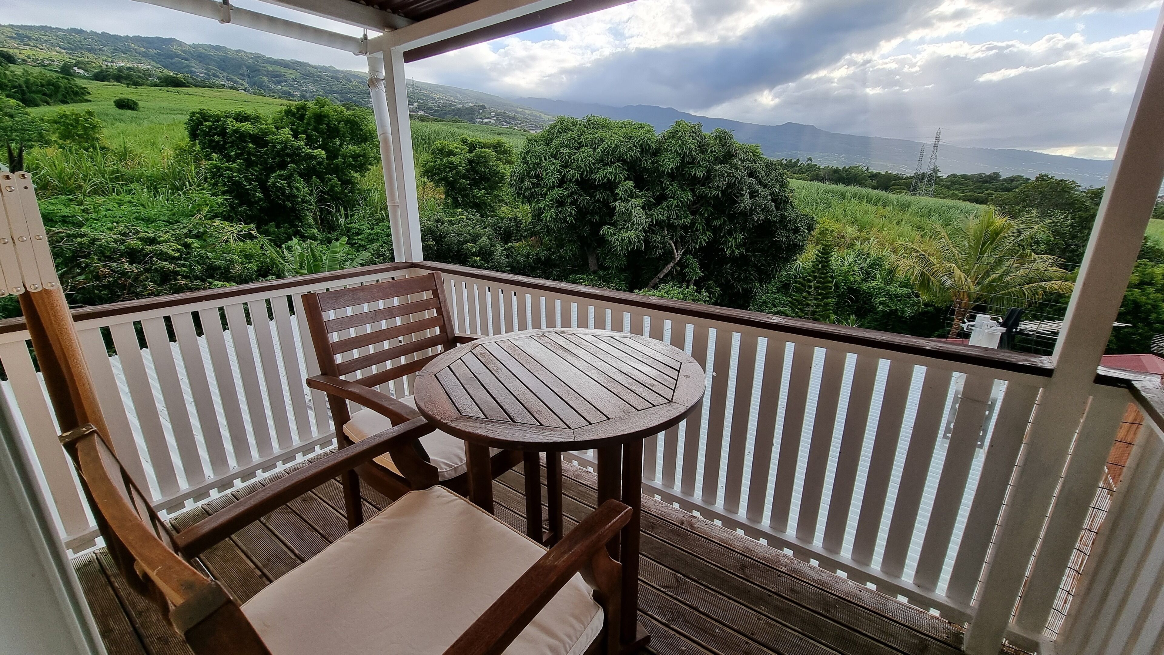 A balcony with a wooden chair and table, offering a view of green hills and distant mountains.