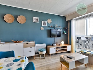 Una sala de estar moderna con una pared azul, un televisor de pantalla plana, un mueble de madera, una mesa de comedor con sillas azules y una ventana con cortinas estampadas.