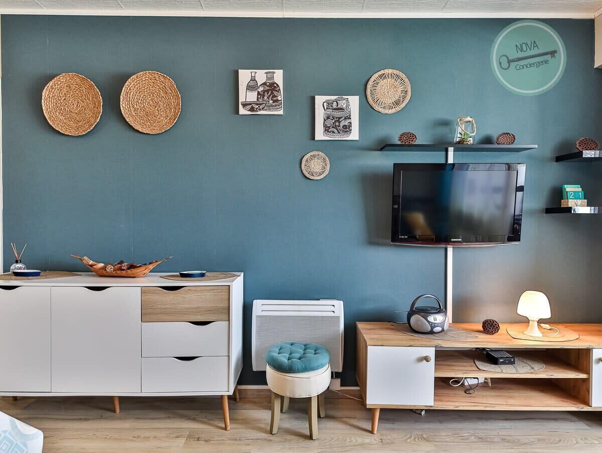 Una sala de estar moderna con un televisor de pantalla plana, un mueble blanco, un otoman azul y platos decorativos tejidos en la pared.