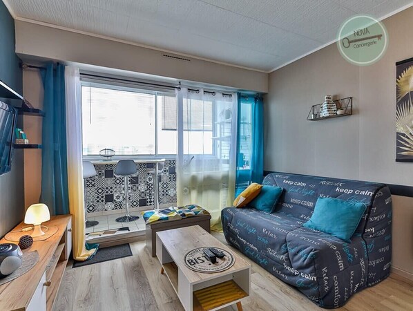 Una sala de estar moderna con un sofá, una mesita de centro y una pequeña mesita auxiliar. Hay una ventana con cortinas y vistas a la ciudad.