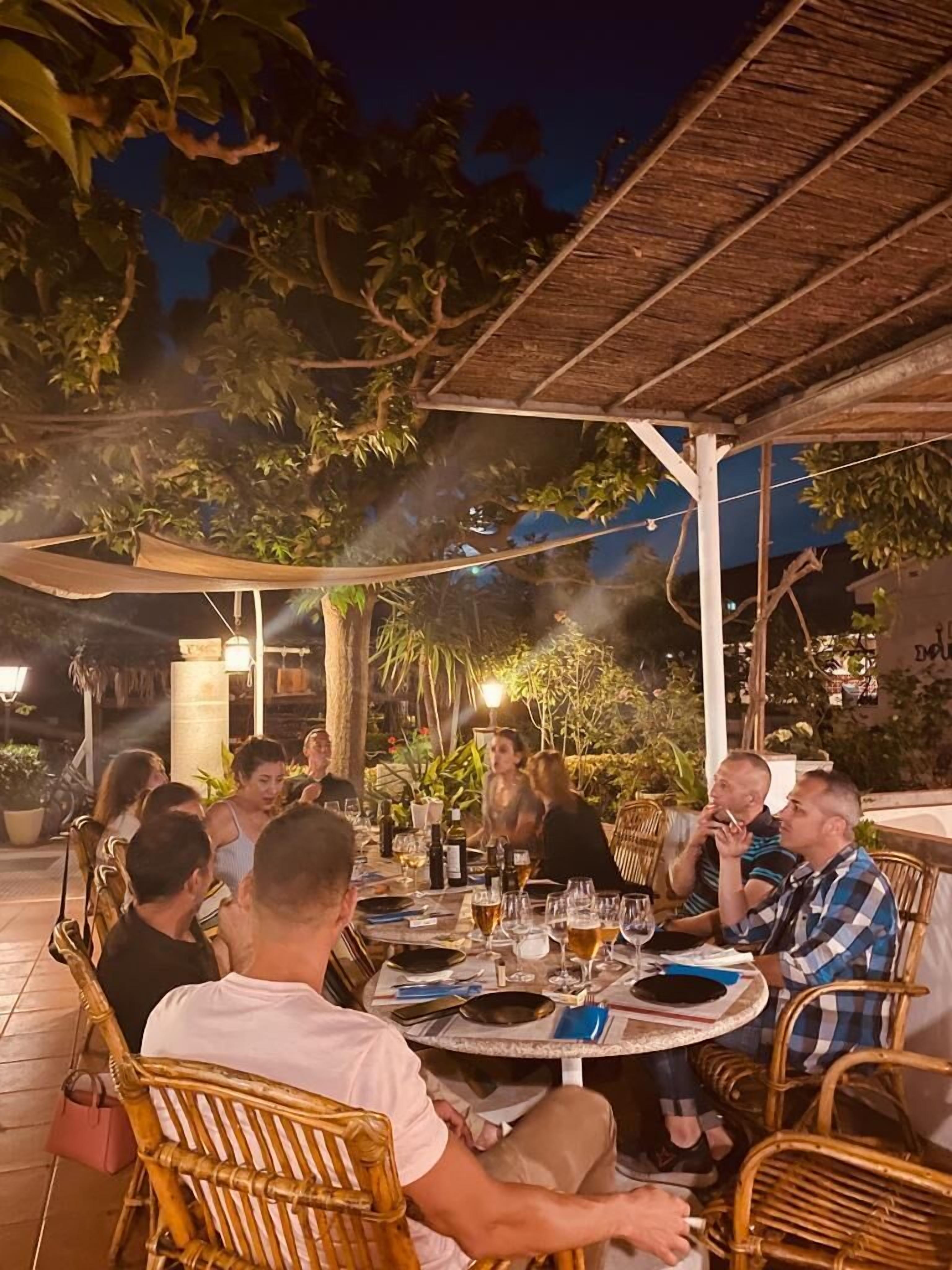 A group of people seated at an outdoor table with drinks, under a thatched roof, surrounded by greenery.