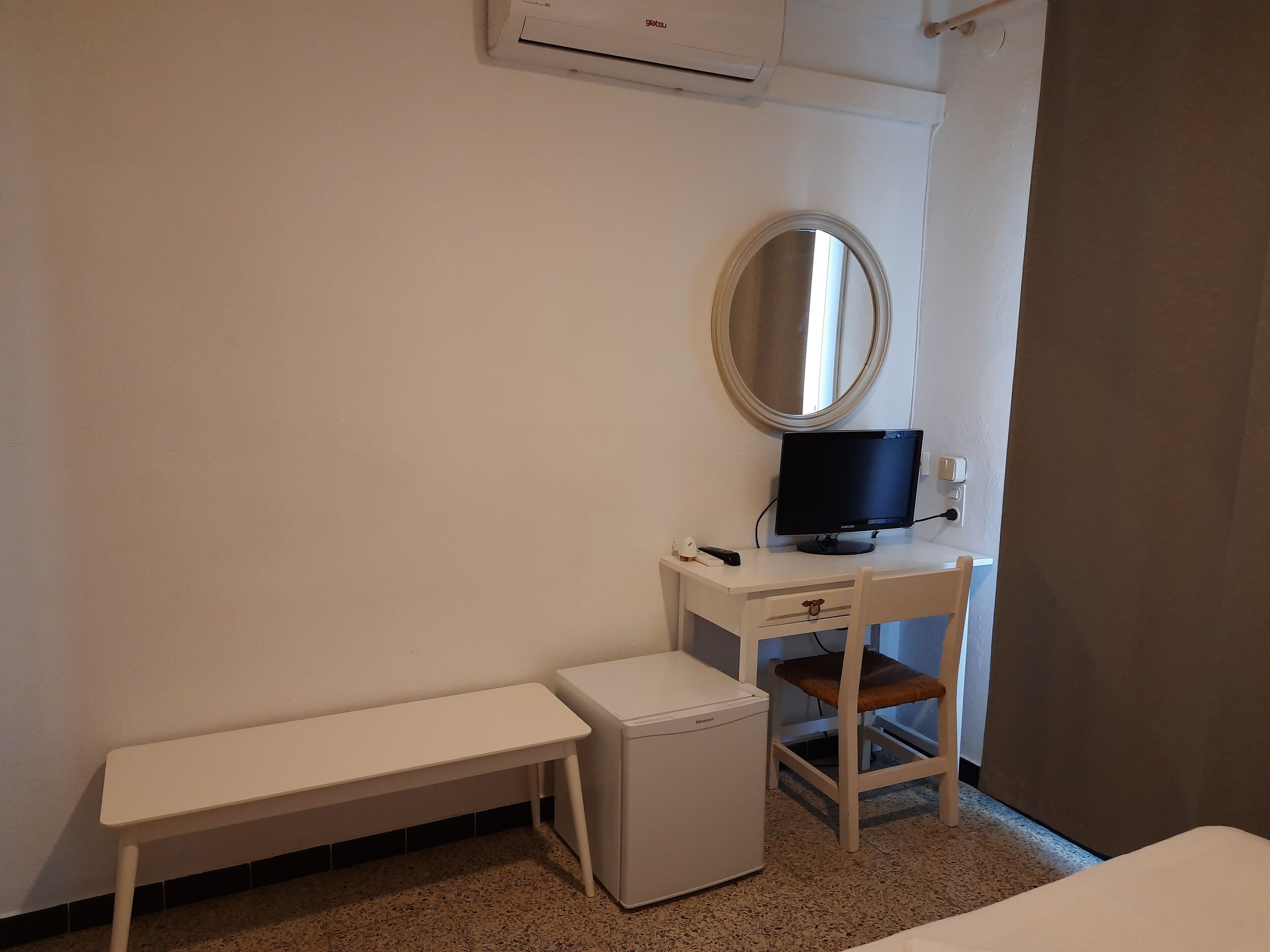 A small, clean room with a white desk, a small white heater, a bench, a mirror, and a television.