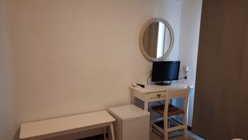 A small, clean room with a white desk, a small white heater, a bench, a mirror, and a television.