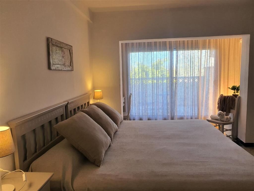 Ein Hotelzimmer mit einem großen Bett, einem Nachttisch mit Lampe, einem Sessel mit Handtuch und einem Fenster mit durchsichtigen Vorhängen.