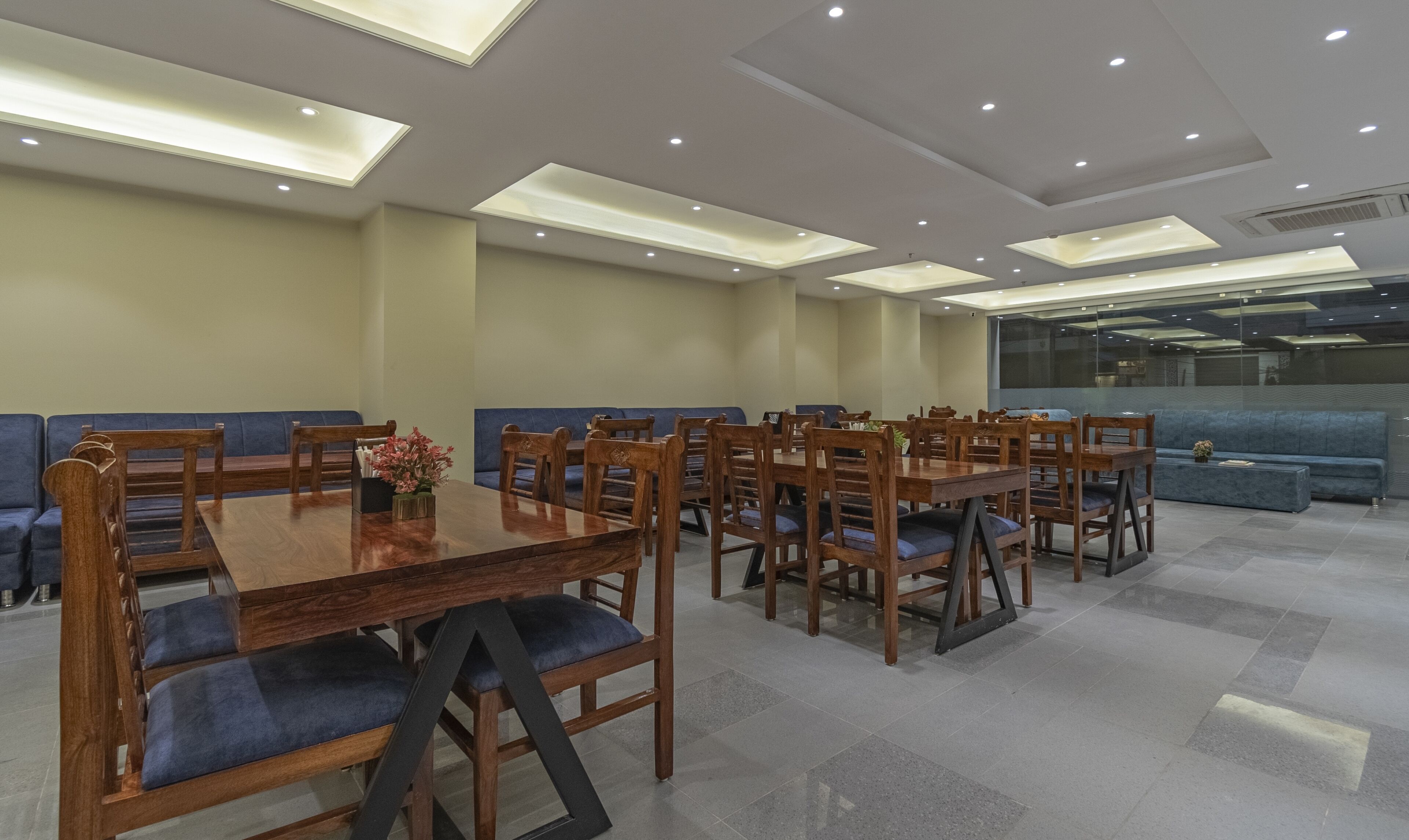 A spacious dining area with wooden tables and chairs, blue cushioned seating, and recessed ceiling lighting.