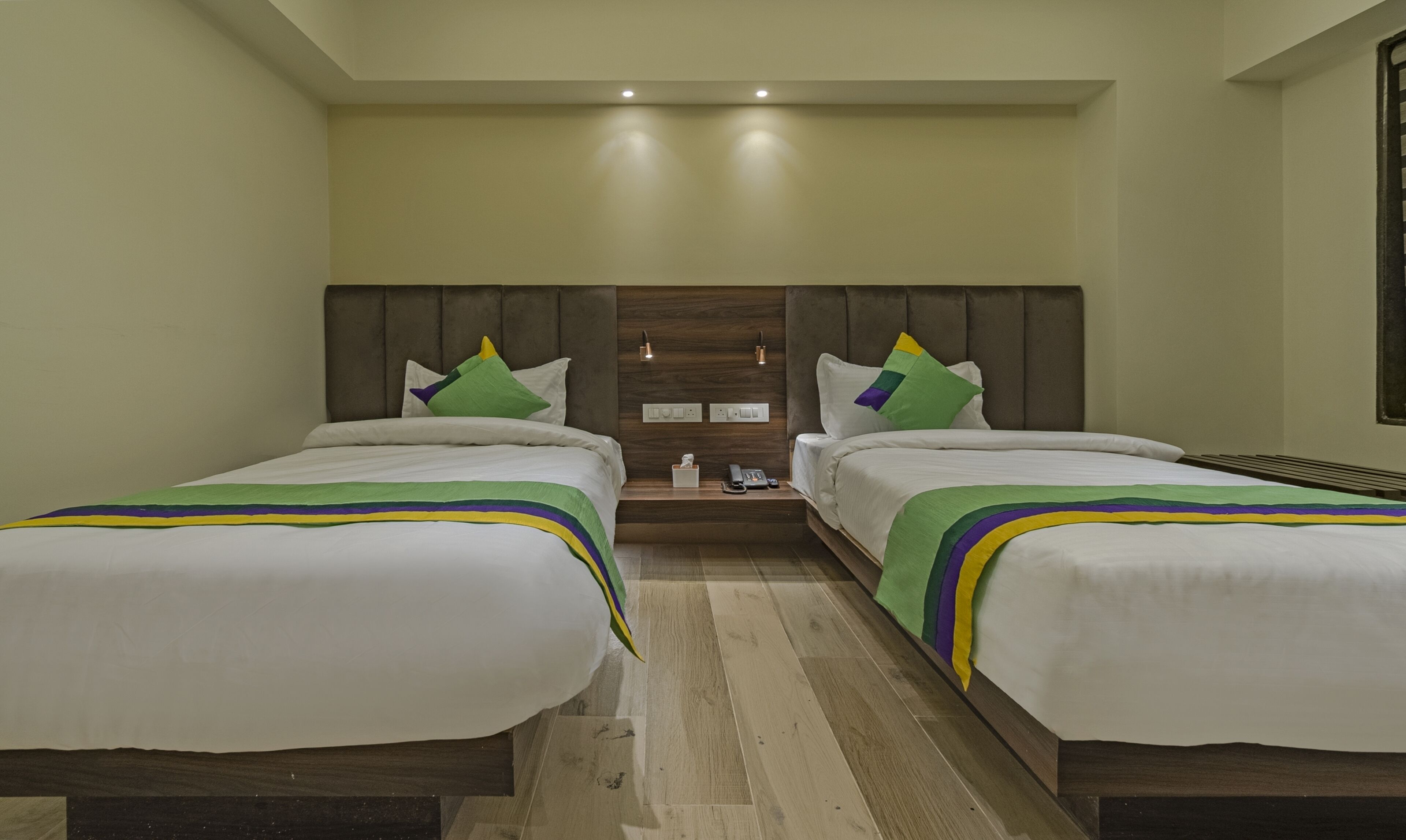 A hotel room with two beds, a wooden headboard, a nightstand, and a wall-mounted light fixture.
