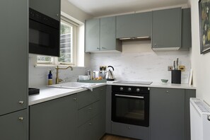 A compact kitchen with a built-in oven, a sink, and a window providing natural light. - Beautifully presented dog friendly - Bea's Cottage (Ampney Crucis, near Cirencester)