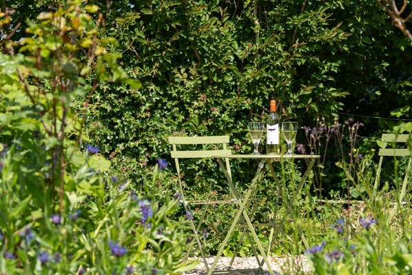 Ett grönt metallbord med en flaska och ett glas vitt vin, omgivet av frodig grönska och lila blommor.