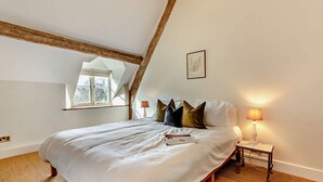 Un dormitorio con techo alto, vigas de madera a la vista, una cama grande con muchas almohadas, una mesita de noche con lámpara y una ventana con vistas a árboles.