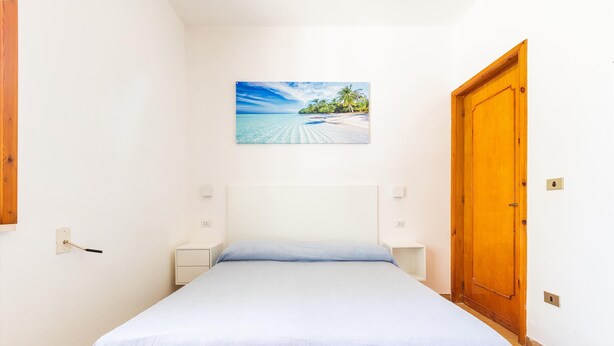 A bedroom with a bed, a wooden door, a nightstand, a wall-mounted light, and a painting of a beach scene. - 3304 Terra del Salento - Mono Costa by Barbarhouse (Porto Cesareo)