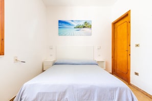 A bedroom with a bed, a wooden door, a nightstand, a wall-mounted light, and a painting of a beach scene. - 3304 Terra del Salento - Mono Costa by Barbarhouse (Porto Cesareo)