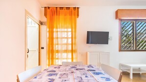 A dining area with a table covered in a patterned tablecloth, a television mounted on the wall, and a window with a metal grille. - 3304 Terra del Salento - Mono Costa by Barbarhouse (Porto Cesareo)