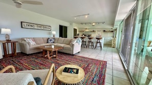 Ein modernes Wohnzimmer mit einer Couch, einem Couchtisch und einem farbenfrohen Teppich. Es gibt eine Küchenzeile mit einer Bar und Hockern, und ein Schild mit der Aufschrift "LIVING THE DREAM".