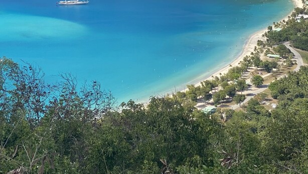 Paesaggio costiero con mare cristallino, una piccola barca a vela e una spiaggia con vegetazione rada.