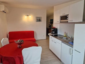 Cozinha compacta com armários brancos, micro-ondas e pia. Há um sofá vermelho e uma mesa de jantar com cadeiras.