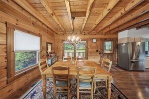 A wooden cabin interior with a dining table, chairs, a chandelier, a kitchen area, and large windows.
