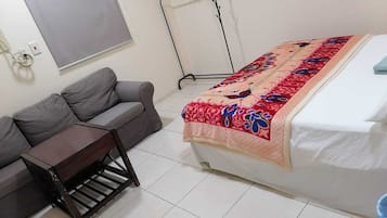 A bedroom with a bed, a grey sofa, a small wooden table, and a standing fan.