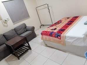 Une chambre à coucher avec un lit, un canapé gris, une petite table en bois et un ventilateur sur pied.