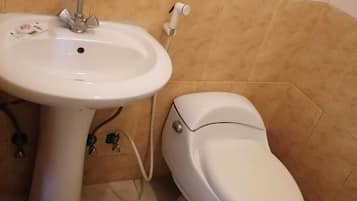 A bathroom with a white sink and toilet, beige tiles on the walls, and a pair of sandals on the floor.