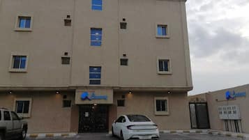 A multi-story building with a beige facade, a central entrance with a black door, and a parking area with a white car.