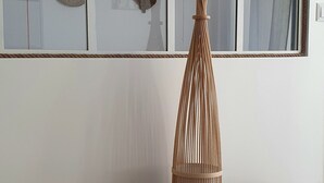 Una habitación con un ventanal grande, una lámpara de pie trenzada, una silla de madera y un otoman tejido.