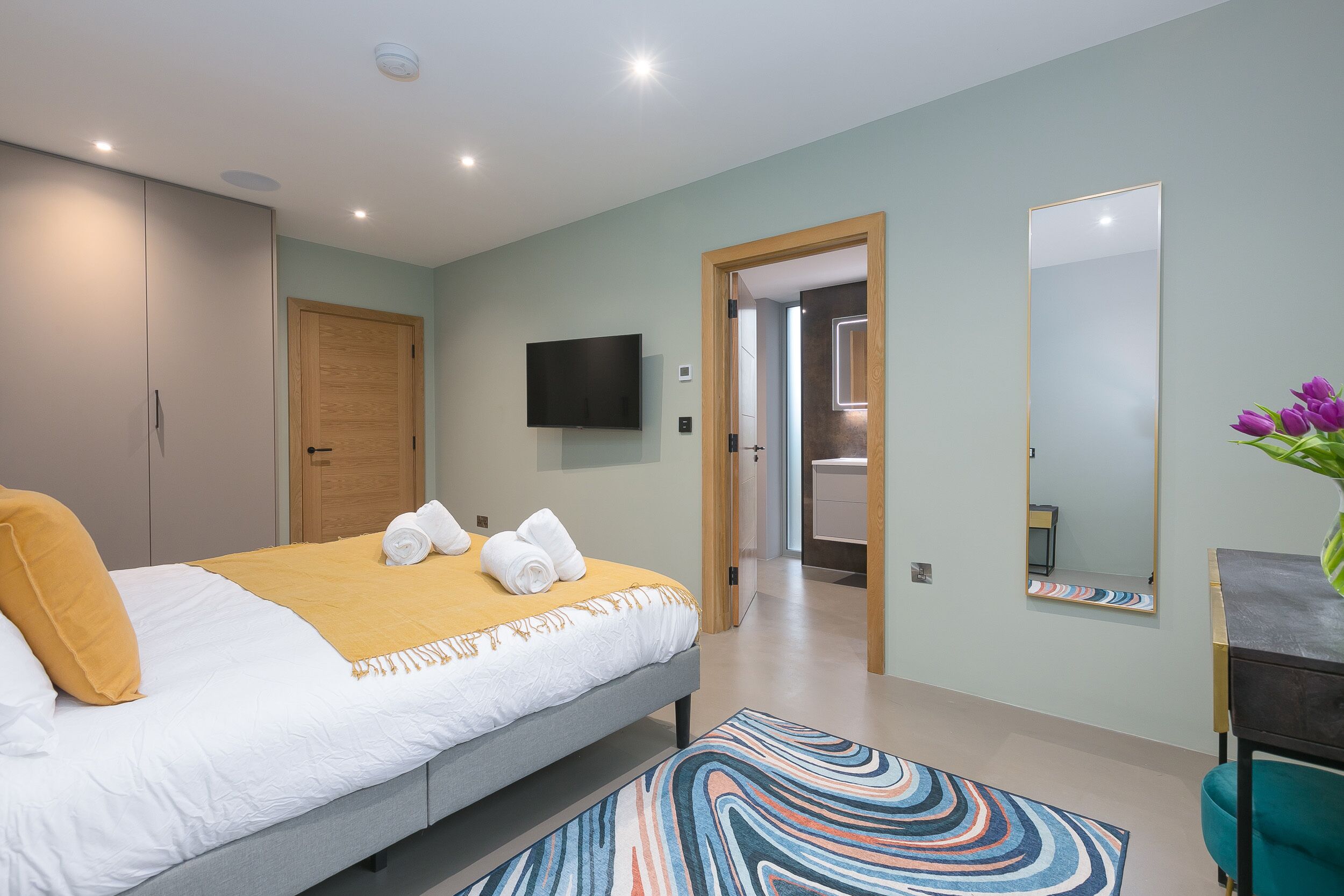 Une chambre moderne avec un lit, une télévision, un miroir et une salle de bain visible à travers une porte ouverte.