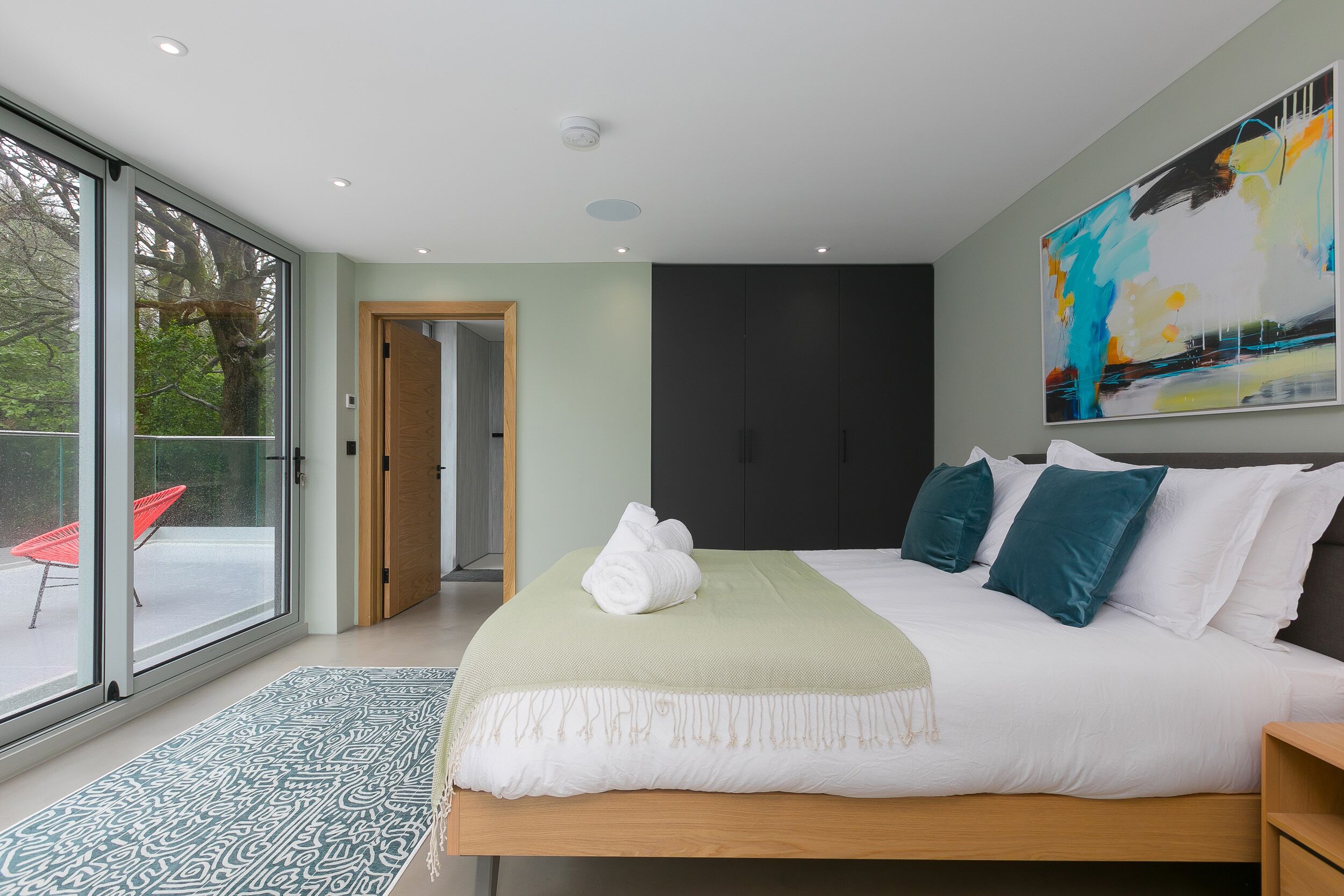 Ein modernes Schlafzimmer mit einem großen Bett, Blick durch Glastüren auf einen Außenbereich und einem großen abstrakten Gemälde an der Wand.