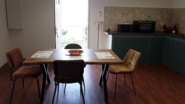 A dining area with a wooden table, four chairs, and a bowl on the table. There is a kitchen area with a microwave and cabinets in the background.