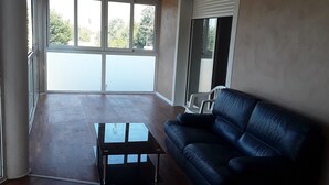 A spacious living room with a black leather sofa, a glass coffee table, and a view of a balcony through large windows.