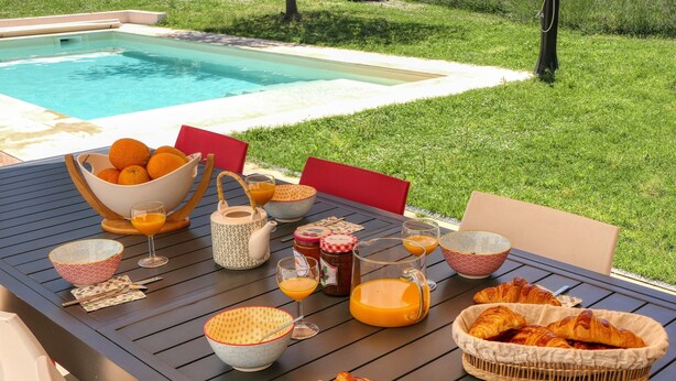 Un buffet de petit-déjeuner avec des croissants, du jus d’orange et divers accompagnements, disposé sur une table près d’une piscine.