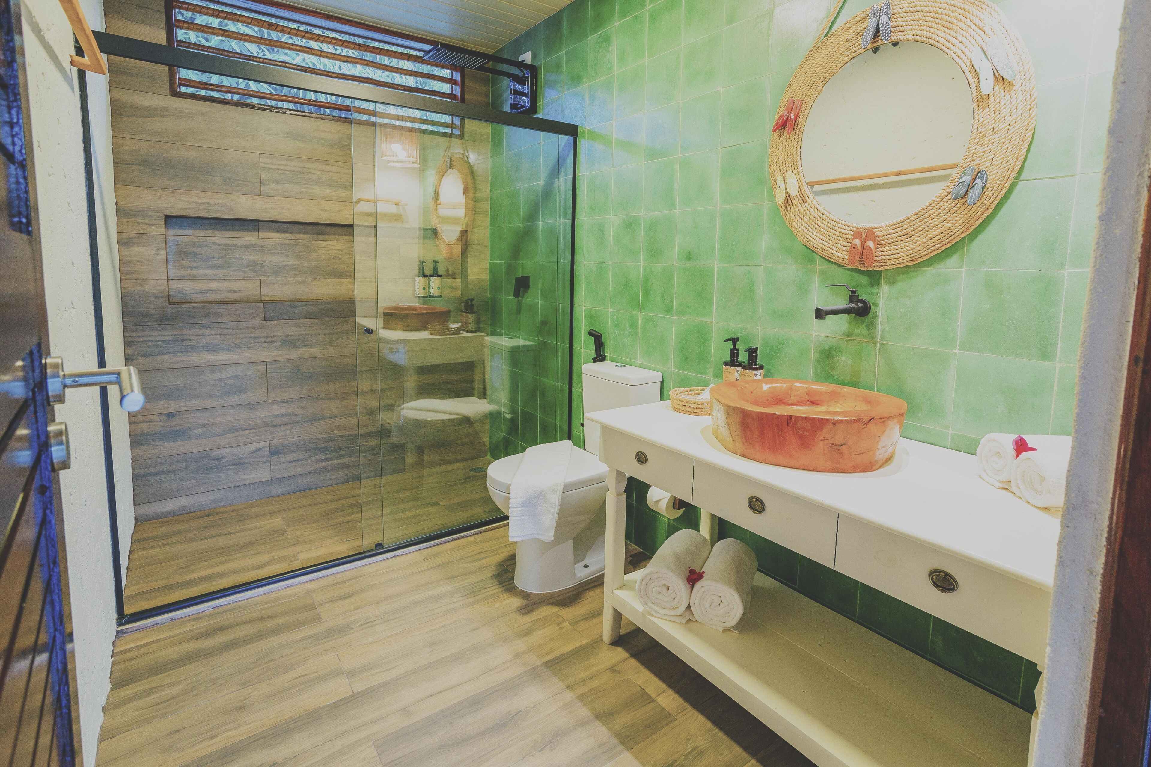 Um banheiro com box de vidro, vaso sanitário branco, pia com cuba de cobre e espelho com moldura trançada.