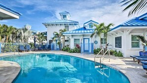 Una piscina con sillas de descanso y palmeras, rodeada de edificios con techos azules y paredes blancas.