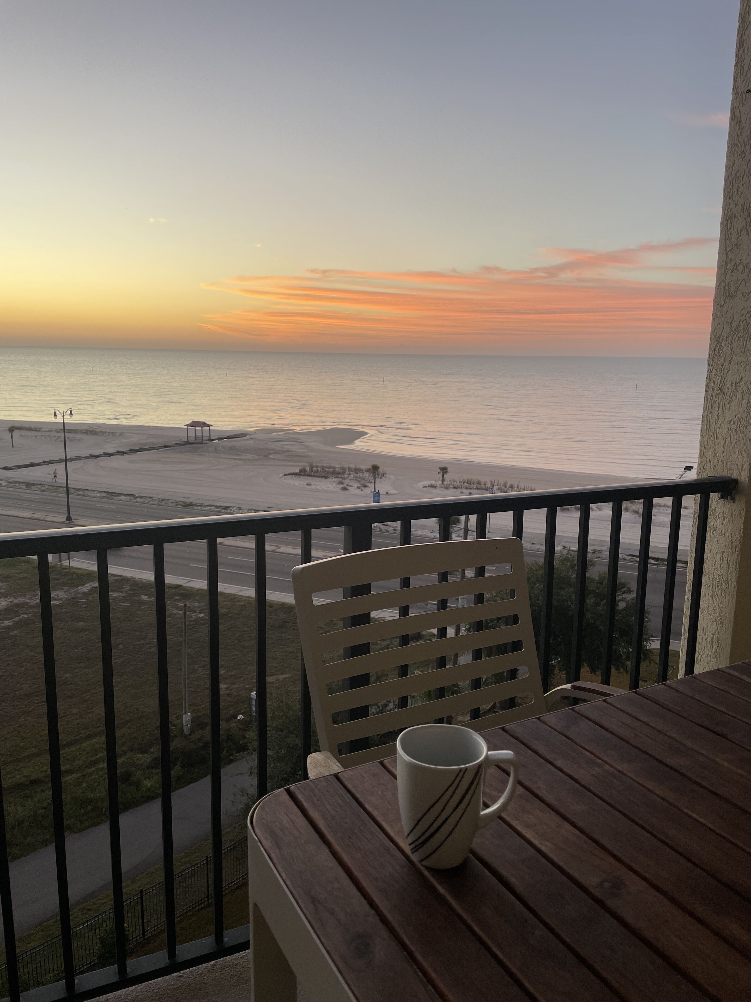 A balcony with a wooden table and a white mug, overlooking a beach and the ocean during sunset.