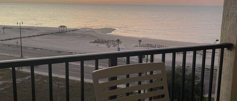 A balcony with a wooden table and a white mug, overlooking a beach and the ocean during sunset.