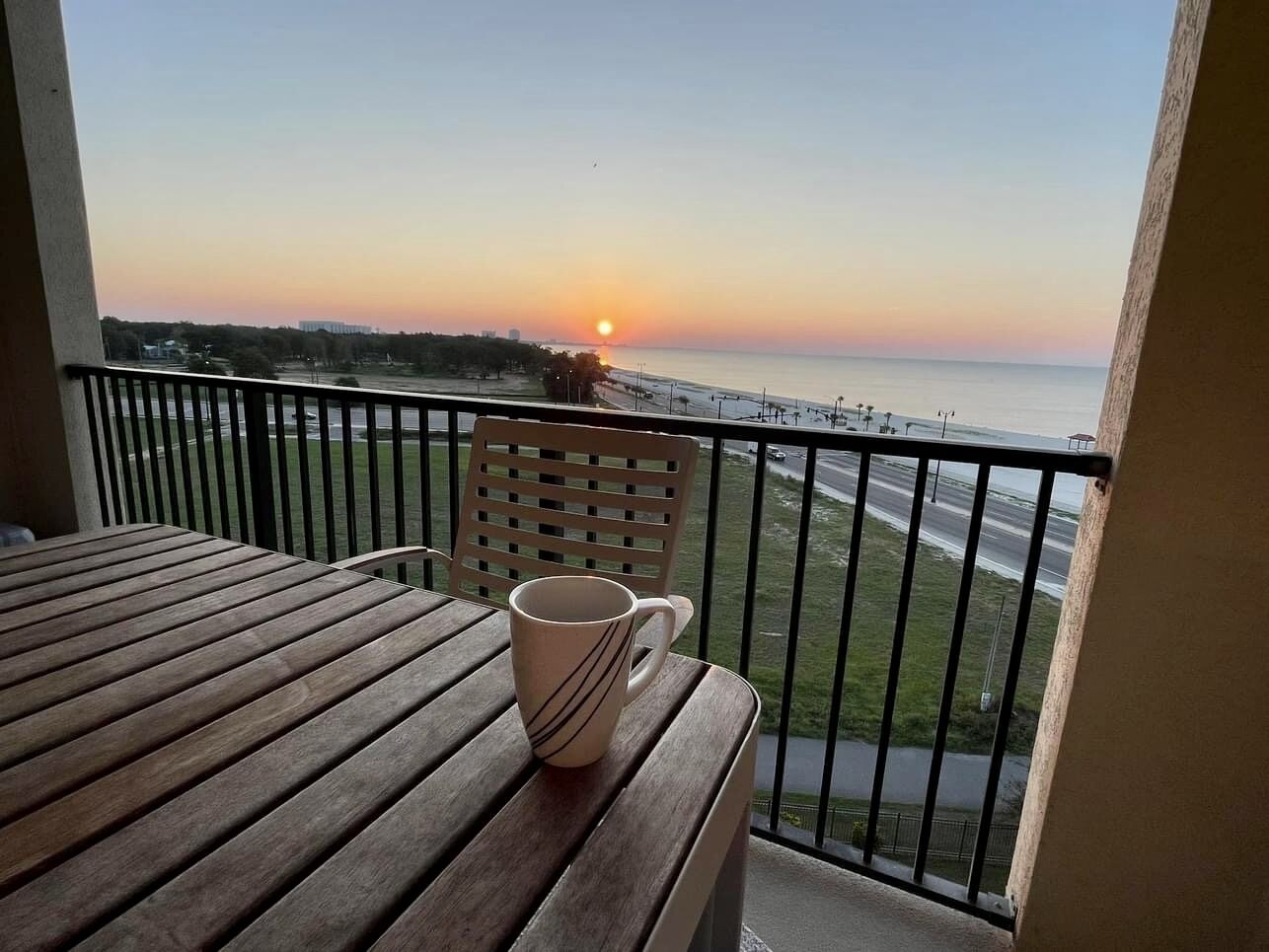 A balcony with a wooden table and a mug, overlooking a sunset over the ocean.