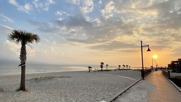 Un sendero de arena con una palmera, un paseo marítimo de madera y un atardecer de fondo.